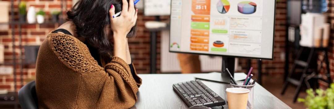Female analyst resting her head in hands, showing exhaustion from office workload. Tired white woman sitting at desk and covering her face, as desktop monitor displays company research information.
