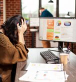 Female analyst resting her head in hands, showing exhaustion from office workload. Tired white woman sitting at desk and covering her face, as desktop monitor displays company research information.