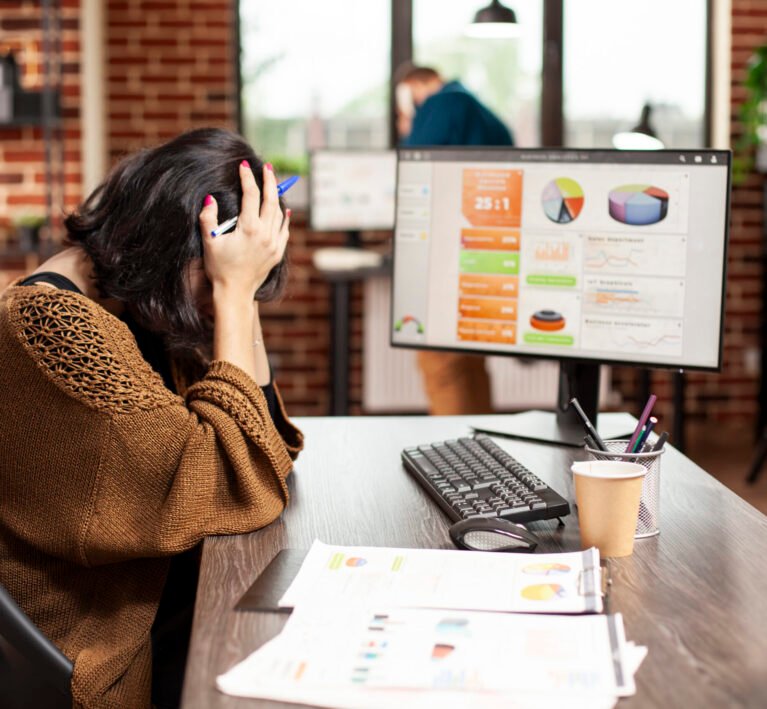 Female analyst resting her head in hands, showing exhaustion from office workload. Tired white woman sitting at desk and covering her face, as desktop monitor displays company research information.